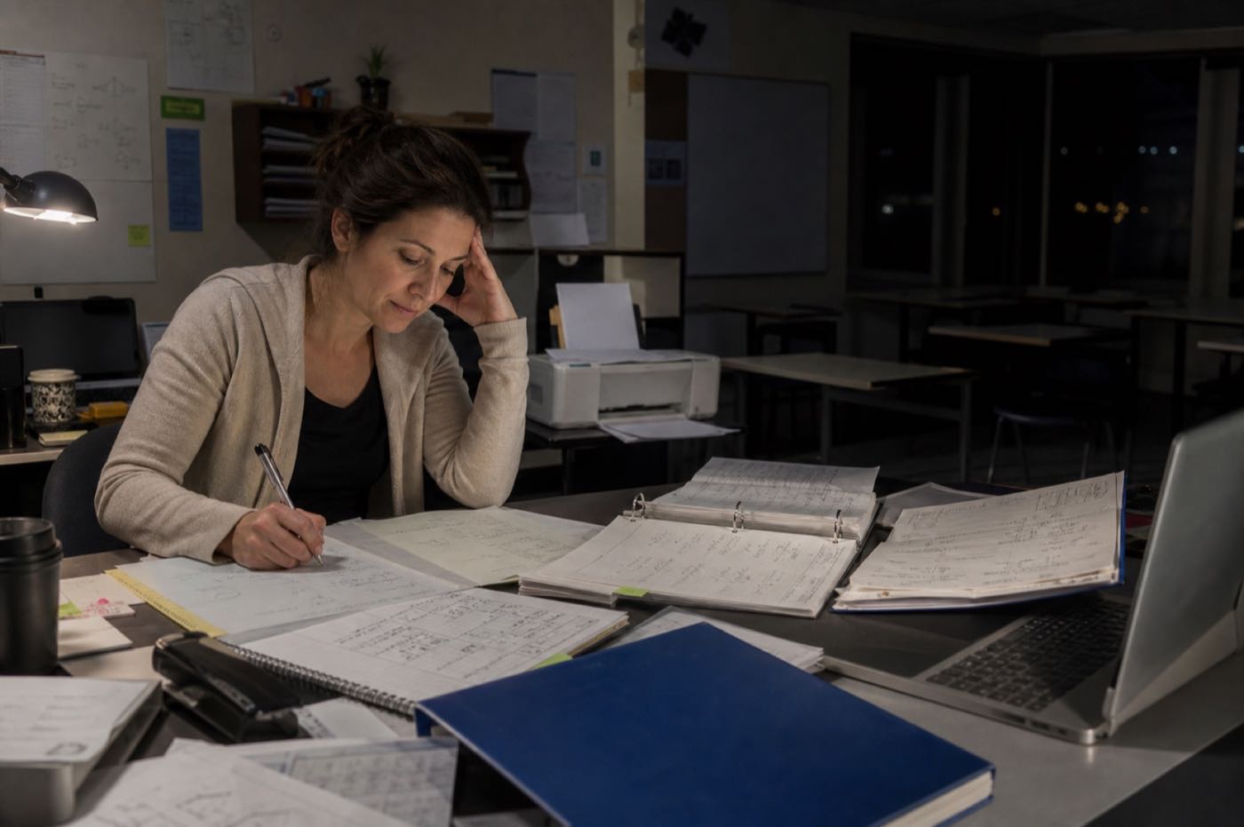 A committed tutor working alone after closing, surrounded by handwritten math work, curriculum binders and printed worksheets while preparing tomorrow's lessons